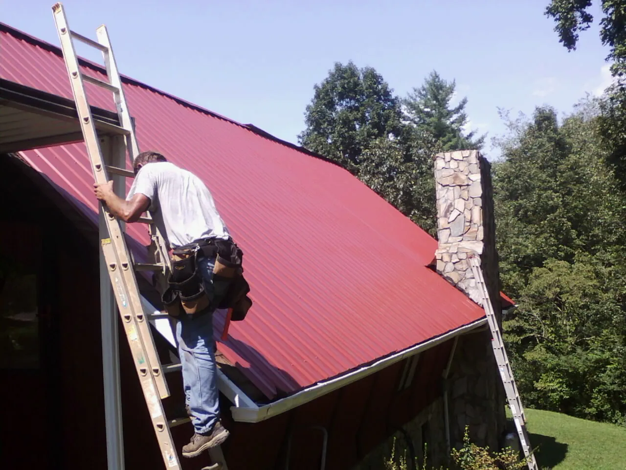 Roofer on a ladder beside a red metal roof and stone chimney — Soffit & Fascia Repair by Roofing Brothers in Honea Path